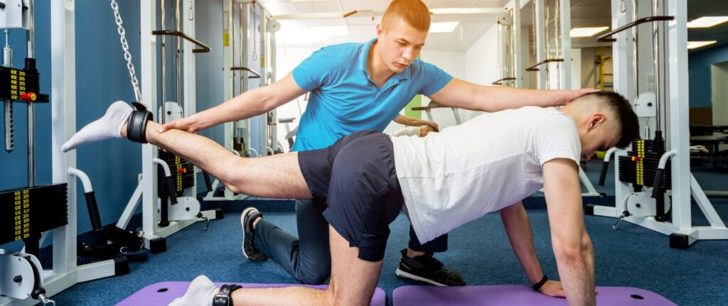 Young man doing exercises on mat under supervision of physiotherapist